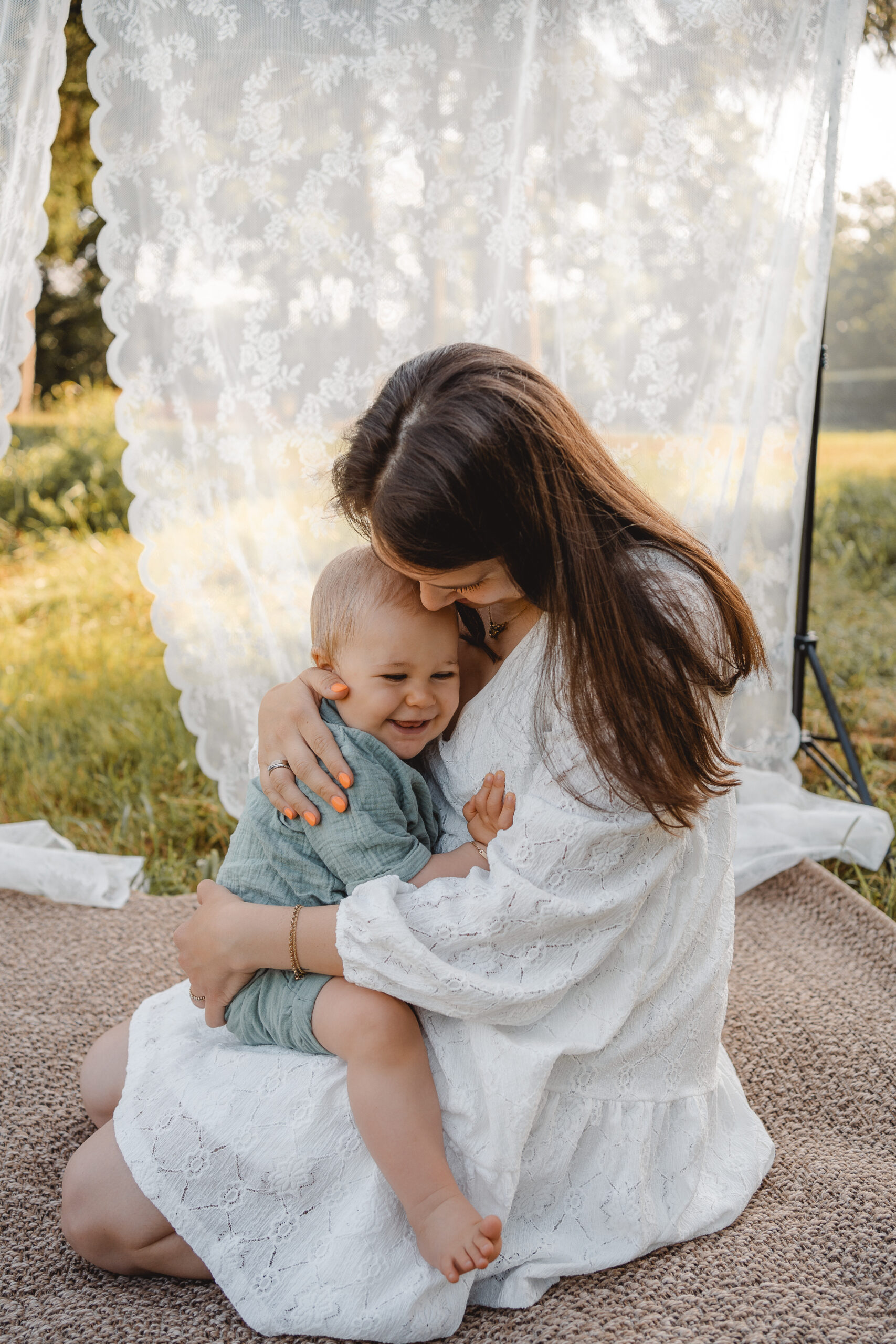 Foto einer Mama mit ihrem kleinem Sohn. Er sitzt auf ihren Schoß und kuschelt sich an ihre Brust, die Mama sieht zu ihm nach unten. Im Hintergrund ist Wiese und ein weißer Spitzenvorhang zu erkennen.