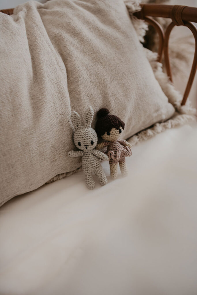 Detailaufname in die Babywiege aus Rattan. Auf einem Kissen liegen zwei gehäkelte Mini-Plüschfiguren. Eine ist ein grauer Hase, die andere eine kleine Puppe mit braunen Haaren, die wie eine Ballerina angezogen ist.