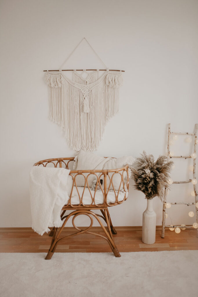 Einblick Fotostudio - Rattan-Babywiege über welcher ein großes weißes Makramee hängt. Daneben steht eine große Vase mit Pampasgras und eine Lichterkette hängt an einer Dekoleiter, die an der weißen Wand lehnt.