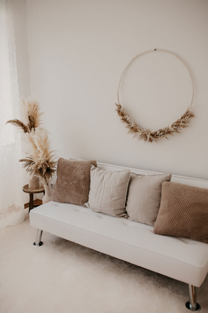 Einblick in mein Tageslichtstudio - Weißes Sofa mit gemütlichen Kissen, darüber hängt ein XXL Trockenblumenkranz und vor dem großen Fenster stehen zwei Vasen mit Pampasgras.