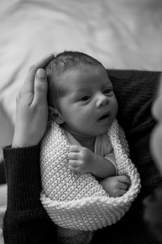 Mama hält ihr Baby auf dem Arm, welches aus der Vogelperspektive fotografiert wurde. Das Baby sieht die Mama mit großen Augen an. Das Baby ist in eine Strickdecke eingewickelt, und die Händchen sehen raus.