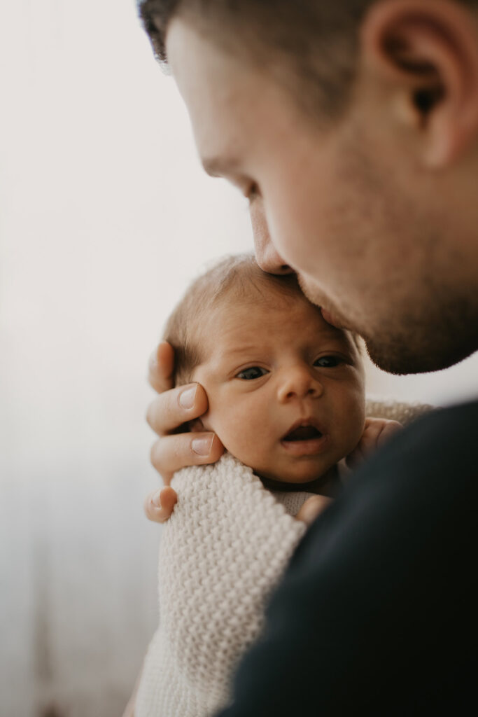 Papa ist leicht von hinten fotografiert, wie er sein Baby an seine Brust hält um ihn ein Küsschen zu geben. Das Baby ist wach und sieht neugierig direkt in die Kamera.