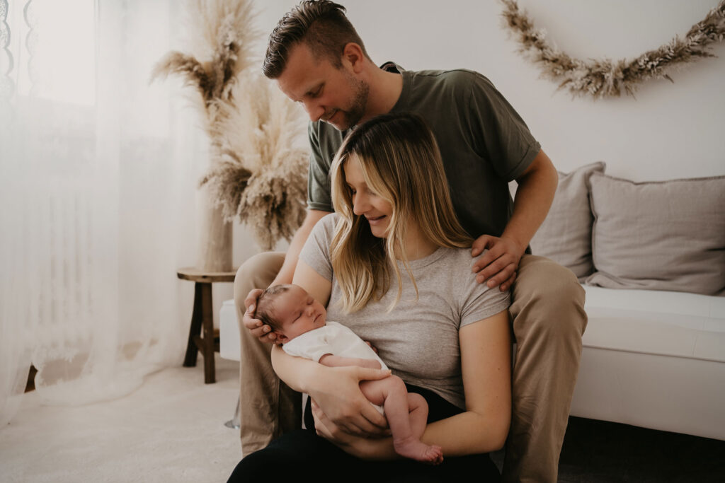 Querformat-Foto einer jungen Familie. Der Papa sitzt auf dem Sofa, die Mama mit dem Baby im Arm zwischen seinen Beinen. Der Papa stützt den Kopf des Babys und beide lächeln es an. Das Baby schläft friedlich im Arm seiner Mama.