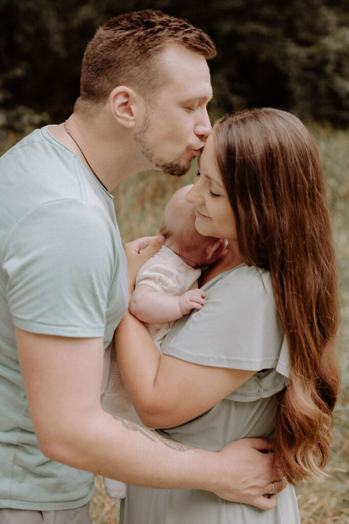 Familie hält Baby zwischen sich, der Papa gibt der Mama einen Kuss auf die Stirn und legt den Arm um ihre Taille.