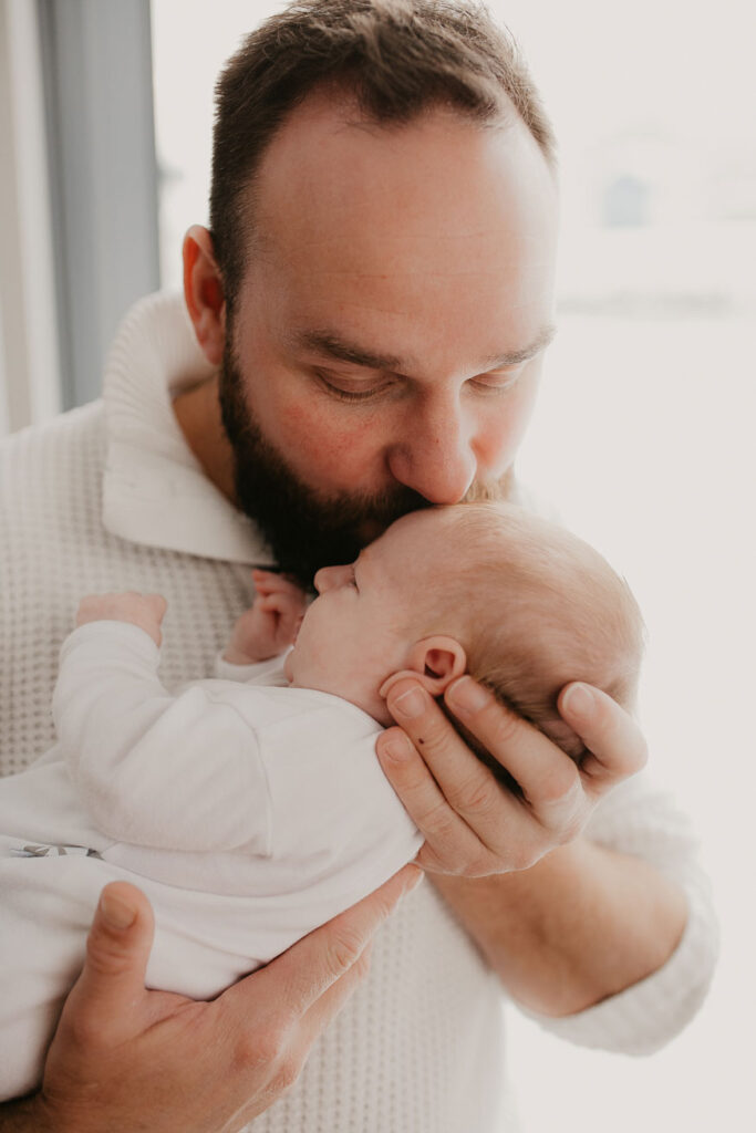 Papa hält das Baby auf dem Arm und stützt seinen Kopf leicht nach oben um ihn ein Küsschen auf die Stirn zu geben. Er steht vor einem bodentiefen Fenster, durch welches die Wintersonne scheint.