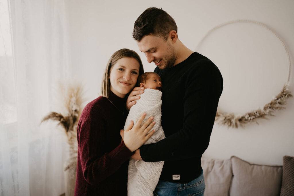 Junge Familie hält ihr Baby zwischen sich auf dem Arm und steht am hellen Tageslichtfenster im Fotostudio. Die Mama lächelt in die Kamera, während der Papa das Baby anlacht.