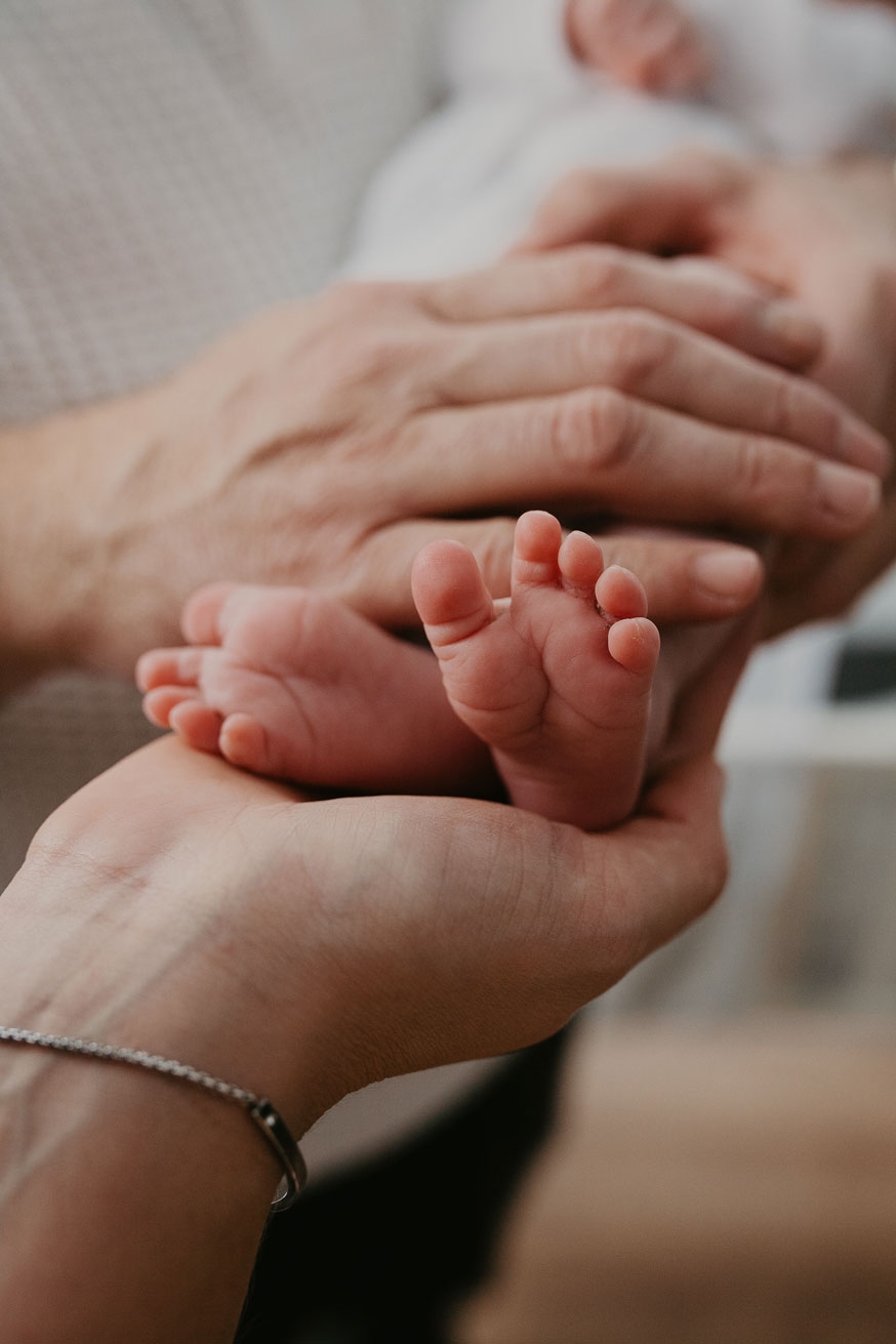 Detailaufnahme von kleinen Babyfüßchen. Die Füßchen befinden sich in der Mitte vom Bild und werden von den Händen der Eltern umrahmend festgehalten.