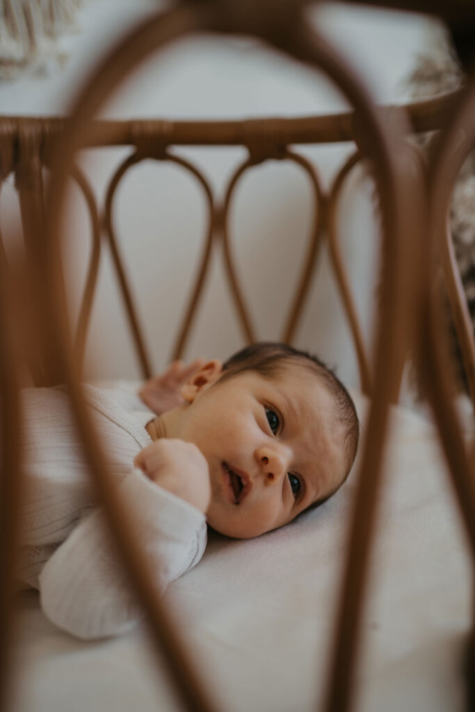 Baby liegt auf dem Rücken in einer Rattanwiege. Das Foto wurde durch die Gitterstäbe aufgenommen und das Baby sieht neugierig in die Kamera.