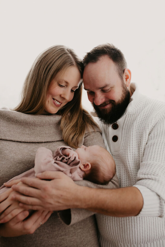 Mama und Papa halten ihr Baby vor sich im Arm und lächeln es an.