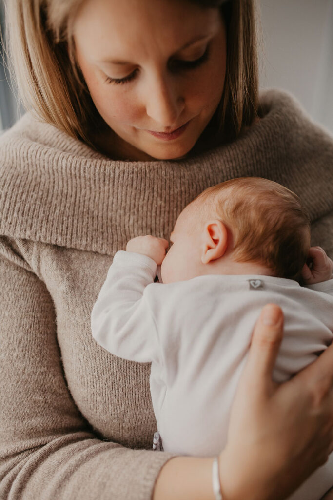 Mama ist frontal zu sehen und hält den Blick nach unten auf ihr Neugeborenes gerichtet, welches sie im Arm hält. Das Baby schläft zufrieden an ihrer Brust.