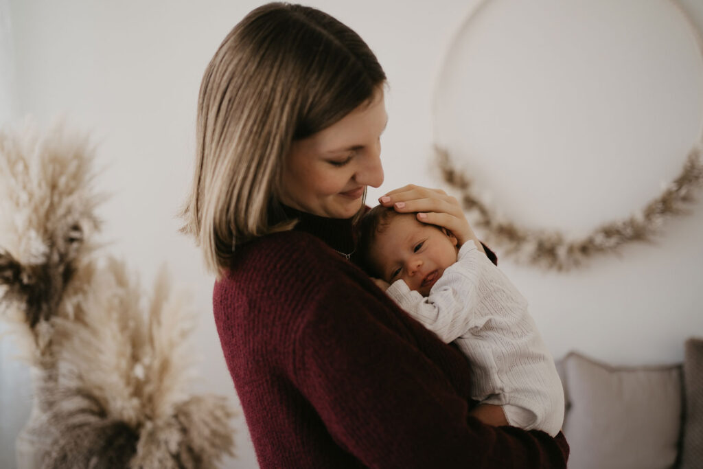 Eine Mama hält ihr Baby in den Armen und streichelt ihm das Köpfchen. Ihr Blick ist liebevoll auf ihr Baby gerichtet, während das Neugeborene neugierig in die Kamera sieht.