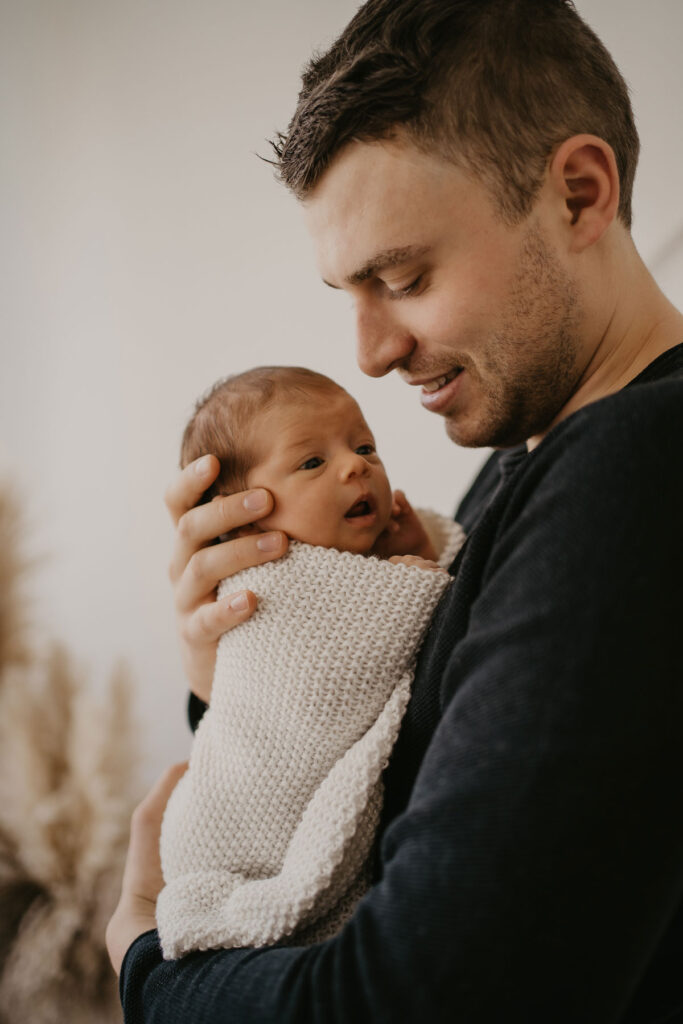 Papa hält Neugeborenes in Decke eingewickelt an seiner Brust. Das Baby ist wach und sieht ihn neugierig an während er es anlächelt.