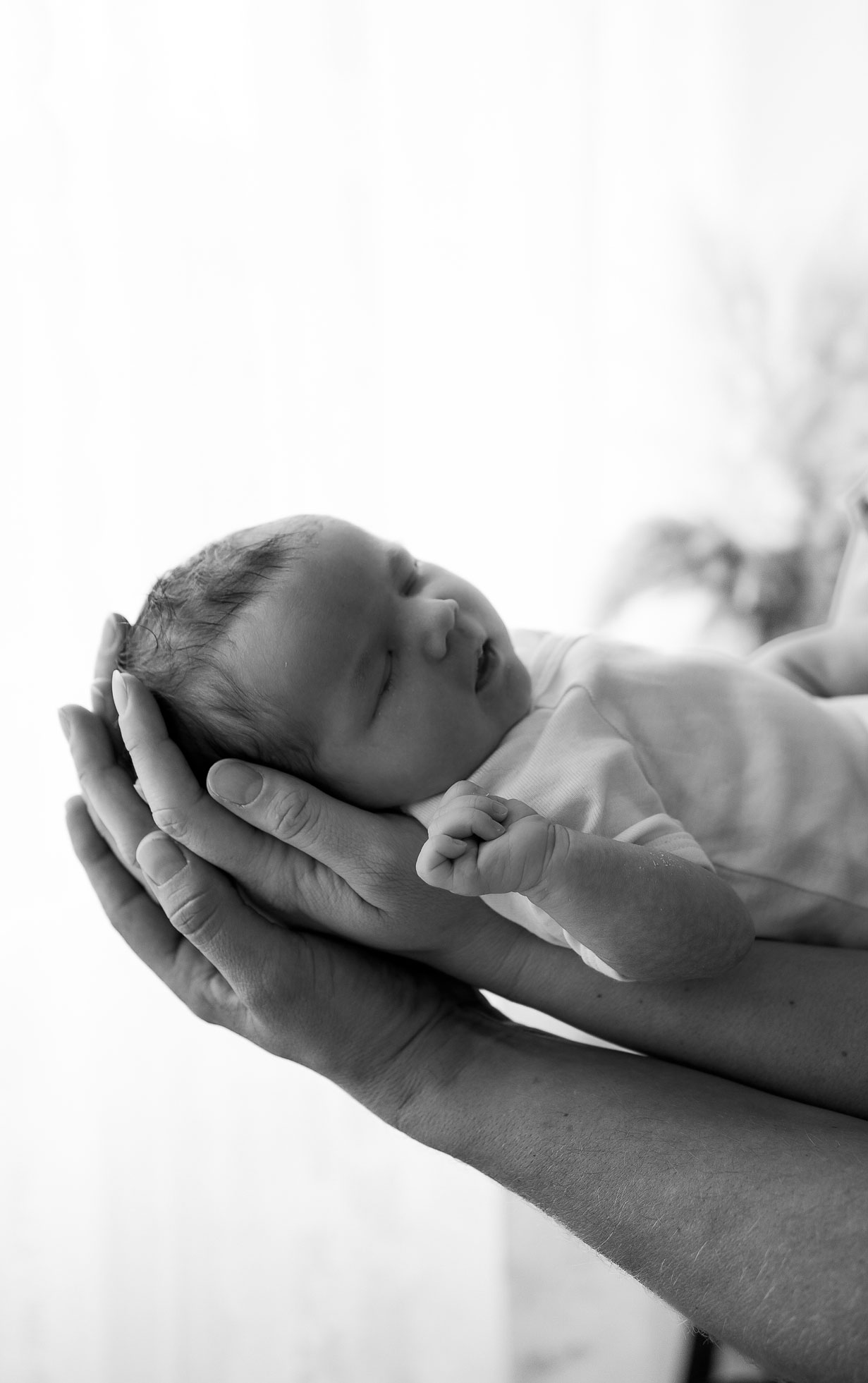 Neugeborenes Baby liegt schlafend in den Armen beider Eltern. Im Hintergrund scheint sanftes Licht durch weiße Spitzenvorhänge.