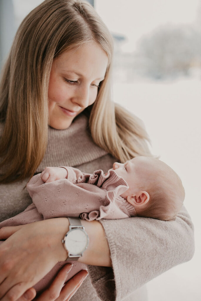 Mama hält ihr Baby auf dem Arm, der Kopf ist dem Baby zugewandt. Sie stehen vor einem bodentiefen Fenster, durch das sanft die Wintersonne strahlt.