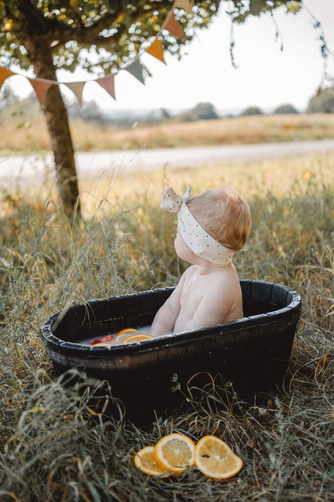 Baby sitzt draußen in einer kleinen Badewanne voller Milch und Früchten. Es ist ein Warmer Sommertag, die Wiese ist gold vom Licht und im Hintergrund ist ein Baum und eine Wimpelkette erkennbar