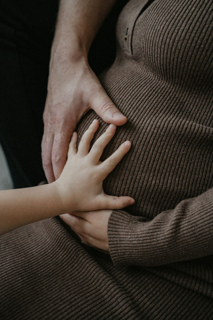 Detailaufnahme eines Babybauches. Der Bauch ist im Fokus des Bilds und wird von den Händen der Mama, des Papas und der großen Schwester gehalten.