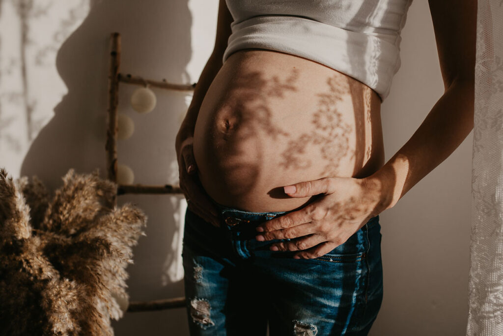 Detailaufnahme eines Babybauches. Das Shirt wurde nach oben gezogen, die Frau hält den Bauch fest. Auf dem Bauch fallen Schatten vom Spitzenvorhang des großen Fensters durch das gerade die Sonne scheint.